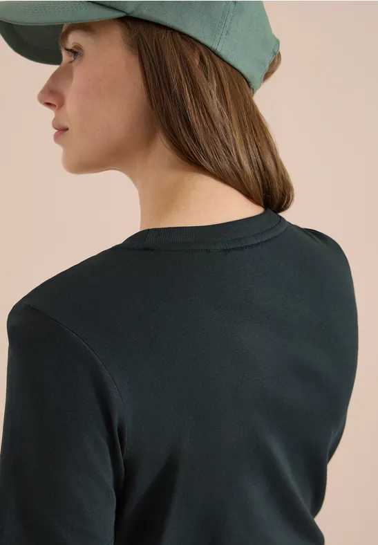 Vue de dos d'une personne portant un t-shirt vert foncé et une casquette vert clair devant un mur beige.