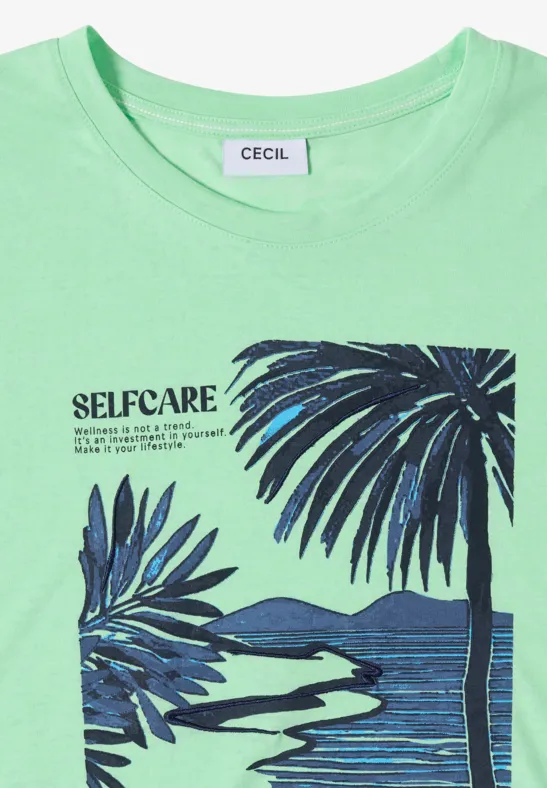 Grünes T-Shirt mit 'SELF-CARE'-Schriftzug und Palmenmotiv vor Wasserlandschaft.