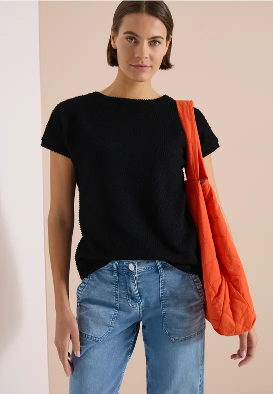 Eine Person trägt ein schwarzes T-Shirt, blaue Jeans und eine orangefarbene gesteppte Umhängetasche.