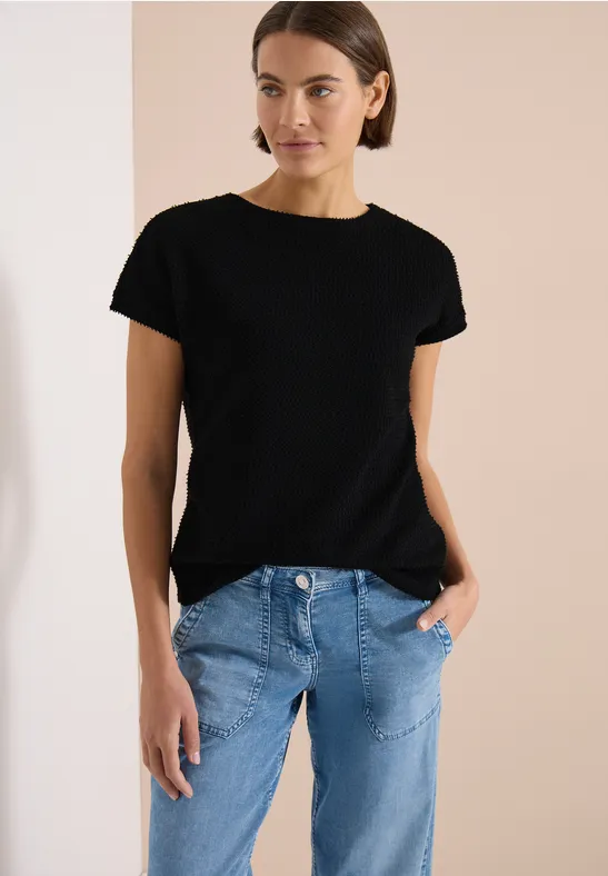 Frau in schwarzem T-Shirt und blauen Jeans vor schlichtem, beigem Hintergrund.