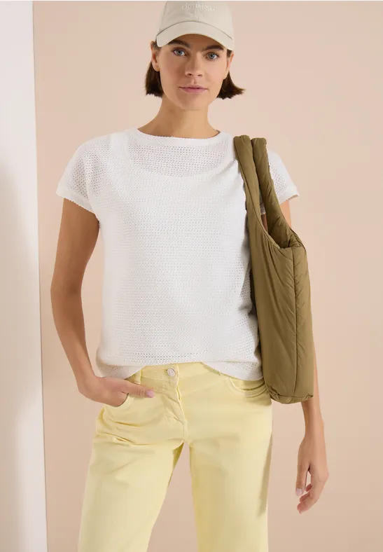 Frau trägt weißes T-Shirt, hellgelbe Jeans und grüne Umhängetasche vor neutralem Hintergrund.