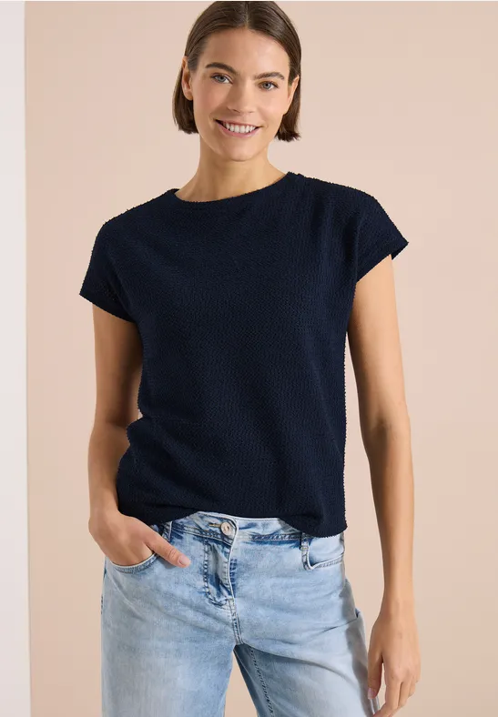 Frau trägt dunkelblaues T-Shirt mit kurzen Ärmeln und helle Jeans vor beigem Hintergrund.