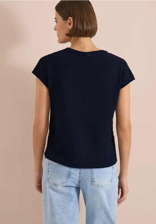 Frau von hinten in dunkelblauem T-Shirt und hellblauen Jeans vor beiger Wand.