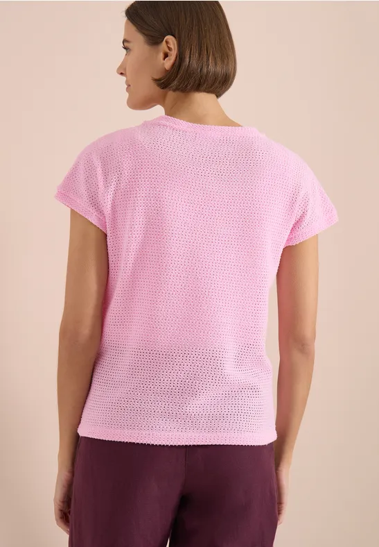 Eine Frau von hinten in rosa Kurzarmshirt und bordeauxroter Hose vor hellem Hintergrund.