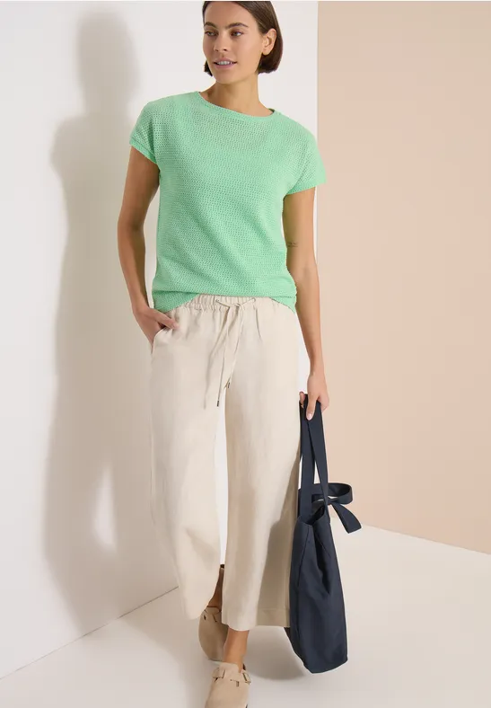 Frau in grünem T-Shirt und weißer, weiter Hose mit Schlitz, trägt eine blaue Tasche vor hellem Hintergrund