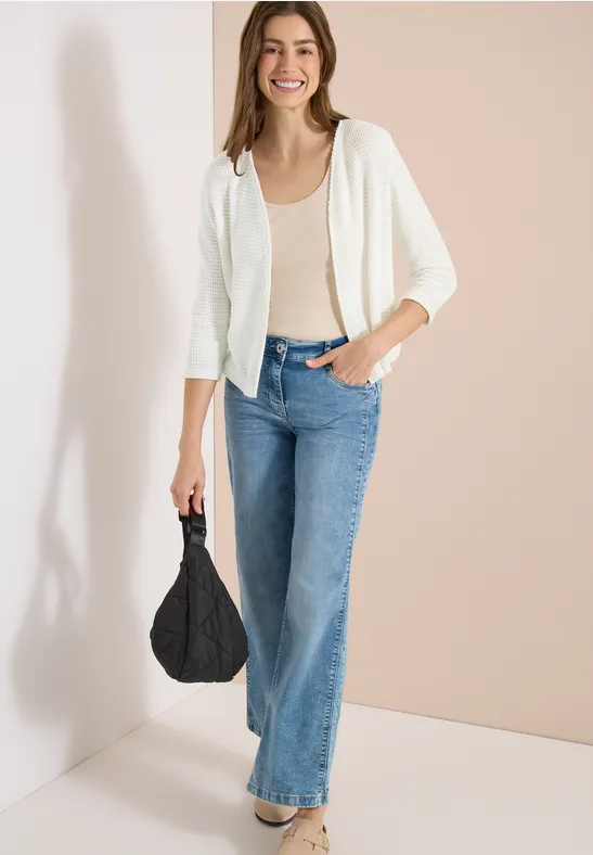 Femme en débardeur beige, gilet blanc et jean bleu, tenant un sac noir devant un mur beige.