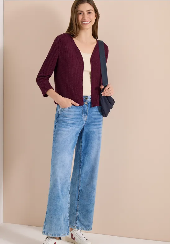 Femme en cardigan violet, top beige et jean large, baskets blanches et sac bleu foncé sur fond uni.