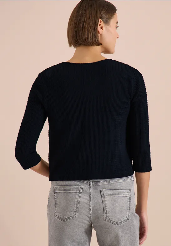 Frau von hinten in schwarzem 3/4-Arm-Shirt und hellgrauer Jeans vor neutralem Hintergrund.