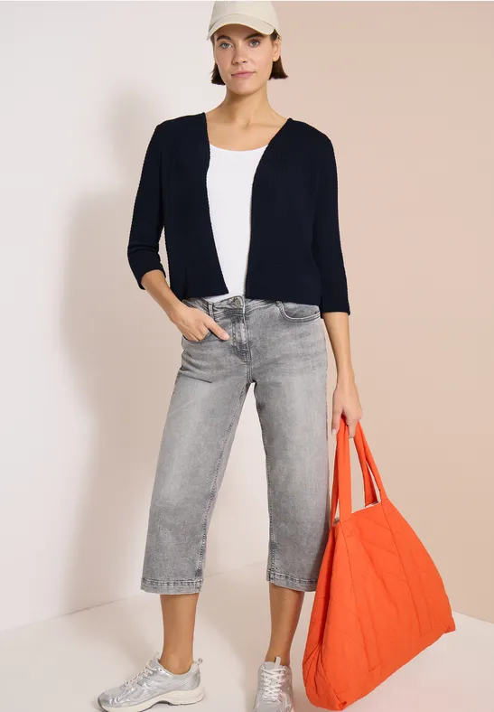 Frau trägt dunkelblauen Cardigan, weißes Top, graue Culotte-Jeans und hält eine große orange Stofftasche.