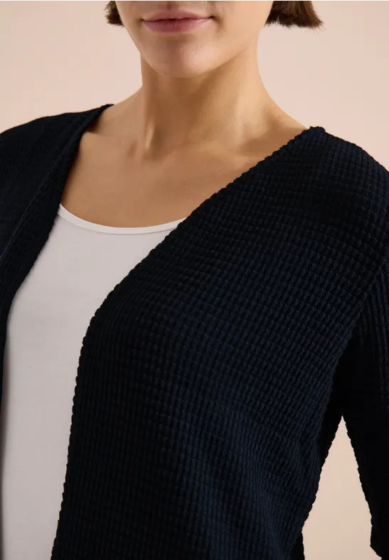 Frau trägt schwarzen Strick-Cardigan über weißem Top, nur Oberkörper sichtbar.
