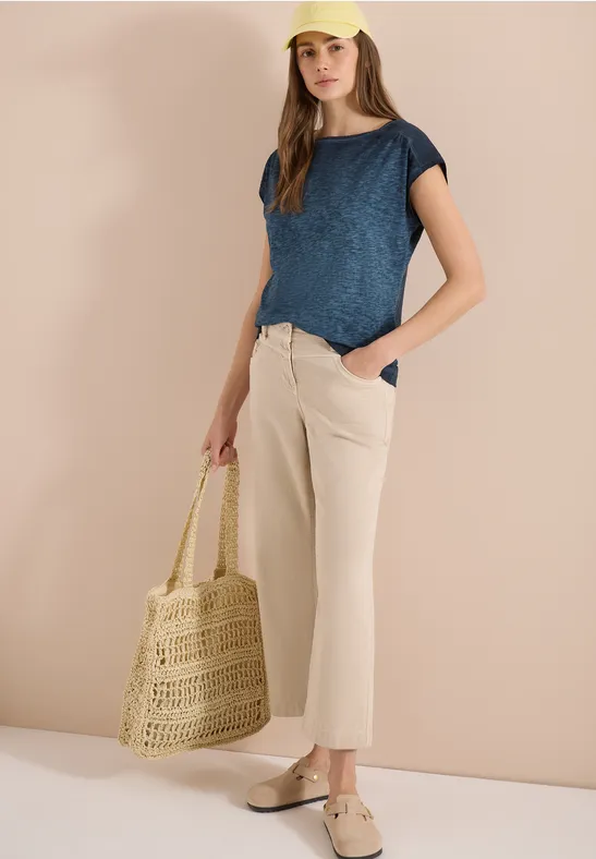 Frau in blauem Shirt, beiger Hose und Espadrilles hält eine Flecht-Tasche vor hellem Hintergrund.