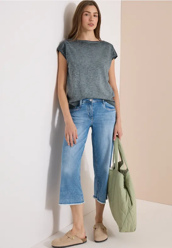 Frau in grauem T-Shirt, hellblauen Cropped-Jeans, beigen Slippern und grünem Steppbeutel vor zweifarbigem Hintergrund.
