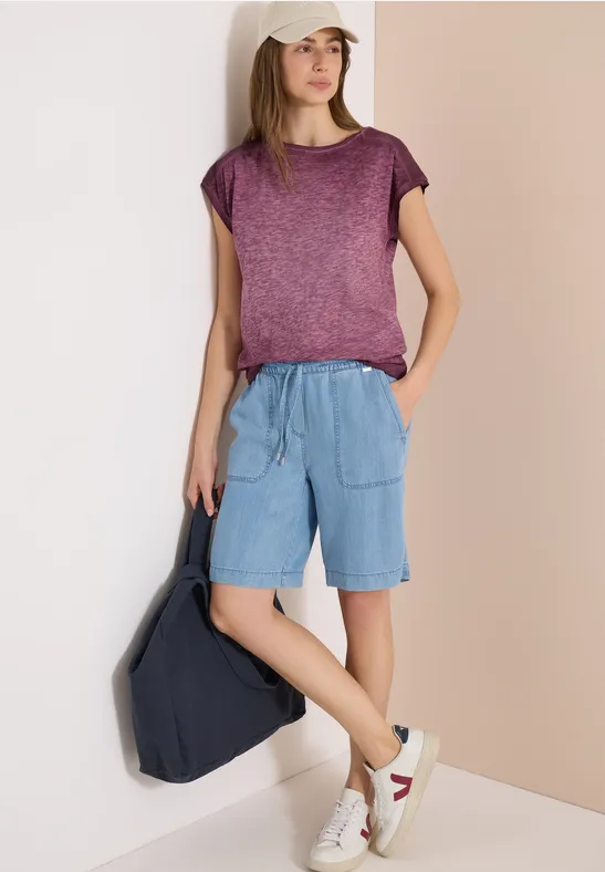 Vrouw met paars t-shirt en lichtblauwe broek, witte sneakers, donkere rugzak, wazig gezicht.