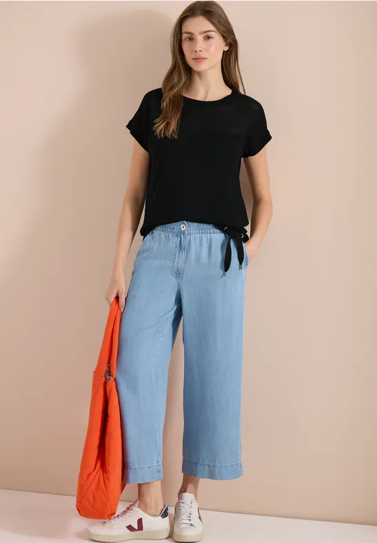 Vrouw met zwart T-shirt, wijde lichtblauwe broek, witte sneakers en oranje tas.
