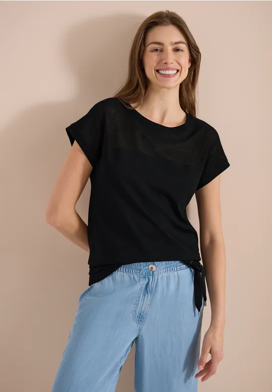 Slanke vrouw met zwart T-shirt en lichte jeans, staand tegen neutrale achtergrond.