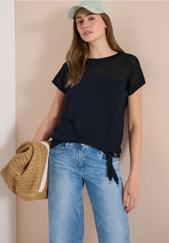 Frau in Jeans, dunkelblauem T-Shirt und Cap hält Strohtasche vor heller Wand