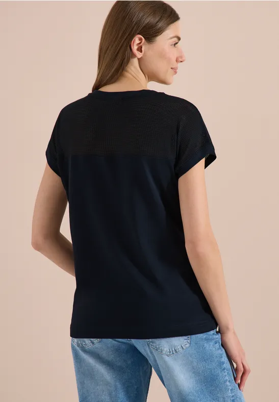 Frau von hinten in dunkelblauem T-Shirt mit kontrastierender Passe und blauen Jeans vor beigem Hintergrund.