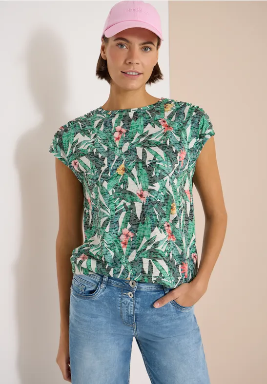 Een persoon draagt een groen t-shirt met tropisch patroon en een jeans, staand voor een lichte achtergrond.