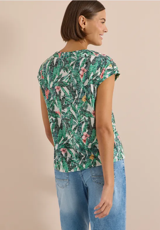 Vrouw van achteren in een groen shirt met tropisch dessin en spijkerbroek voor een neutrale achtergrond.