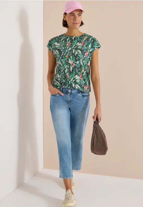 Vrouw in groen tropenprint T-shirt, lichtblauwe cropped jeans, witte sneakers en een bruine tas.
