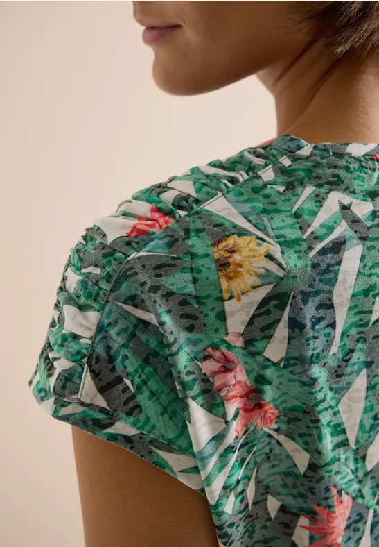 Close-up van een schouder in een groen tropisch shirt met bloemen- en bladpatroon.