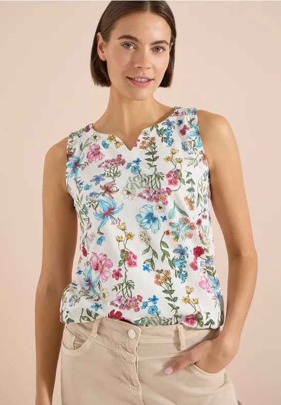 Eine Frau trägt ein ärmelloses, weißes Top mit buntem Blumenmuster und steht entspannt mit Hand in der Tasche.