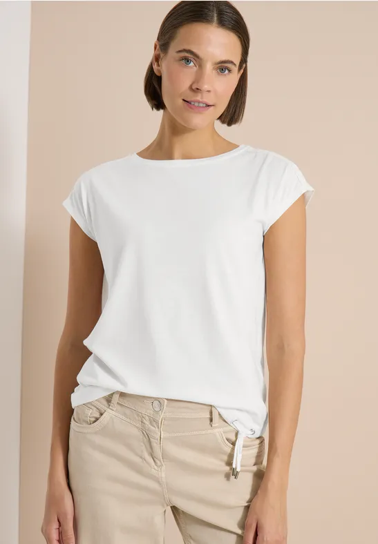 Eine Frau trägt ein weißes T-Shirt und beige Hose vor neutralem Hintergrund.