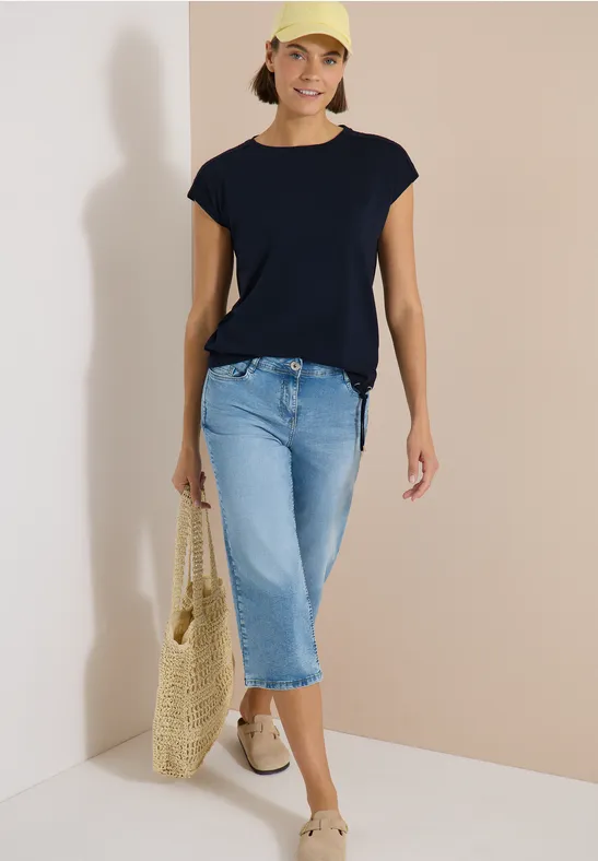 Frau in dunkelblauem T-Shirt, hellblauen Capri-Jeans, hellen Schuhen und gelber Netz-Tasche vor neutralem Hintergrund.