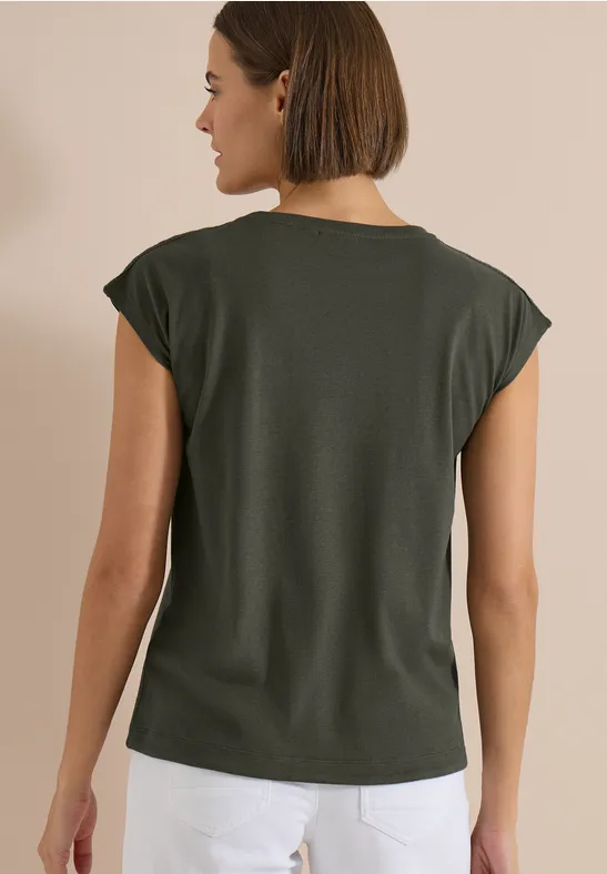 Frau von hinten in dunkelgrünem T-Shirt mit umgeschlagenen Ärmeln und weißer Hose vor beigem Hintergrund.