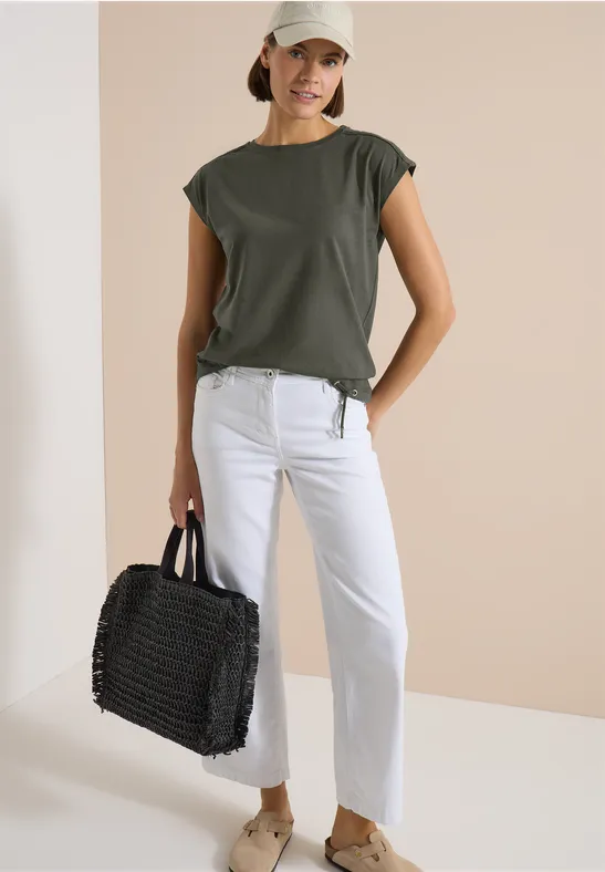 Frau in khakigrünem T-Shirt, weißen Jeans und flachen Sandalen, trägt eine schwarze geflochtene Tasche vor neutralem Hintergrund.