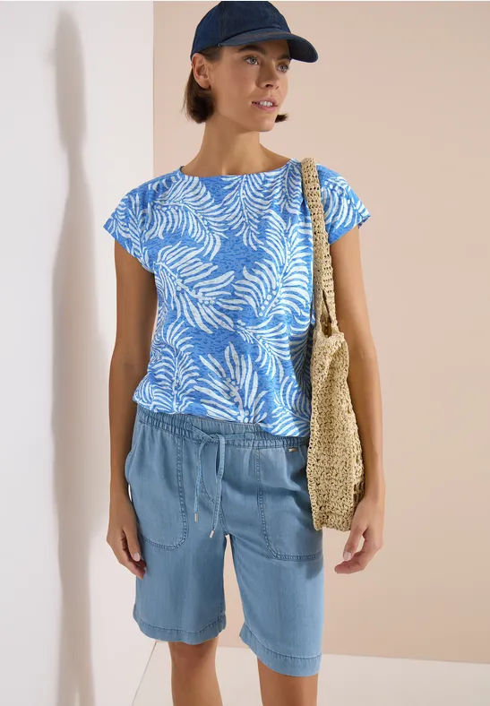 Person mit blau-weiß gemustertem Shirt, Jeans-Shorts, Strohtasche und Cap vor beigem Hintergrund.