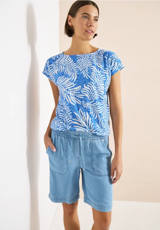 Frau trägt blaues Blattmuster-Shirt und helle Jeans-Bermudashorts vor hellem Hintergrund.