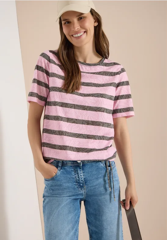 Frau trägt rosa-schwarz gestreiftes T-Shirt und blaue Jeans, steht lässig mit Hand in Tasche.