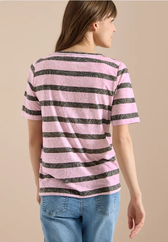 Eine Frau in rosa-braun gestreiftem T-Shirt und Jeans, von hinten vor beiger Wand stehend.