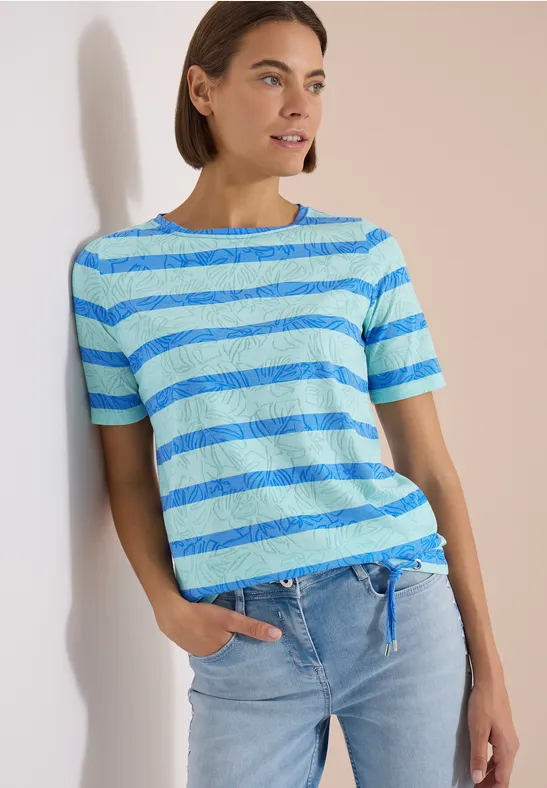 Femme appuyée sur un mur, portant un t-shirt rayé bleu-vert et un jean clair.
