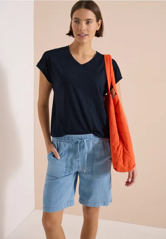 Frau in schwarzem T-Shirt, hellen Jeans-Shorts und rotem Stoffbeutel vor neutralem Hintergrund.
