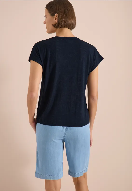 Eine Frau von hinten, trägt ein dunkelblaues T-Shirt und hellblaue knielange Shorts vor neutralem Hintergrund.