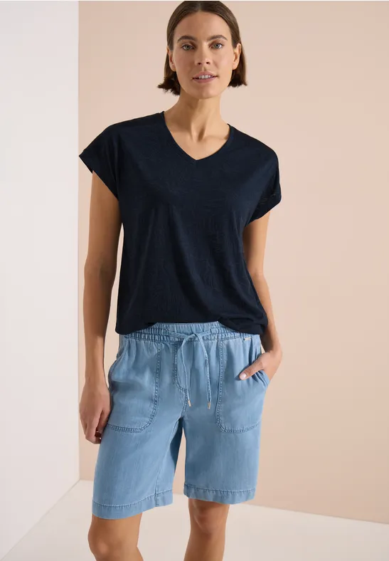 Frau in schwarzem V-Ausschnitt-Shirt und hellblauen Jeans-Shorts mit Taschen vor neutralem Hintergrund.