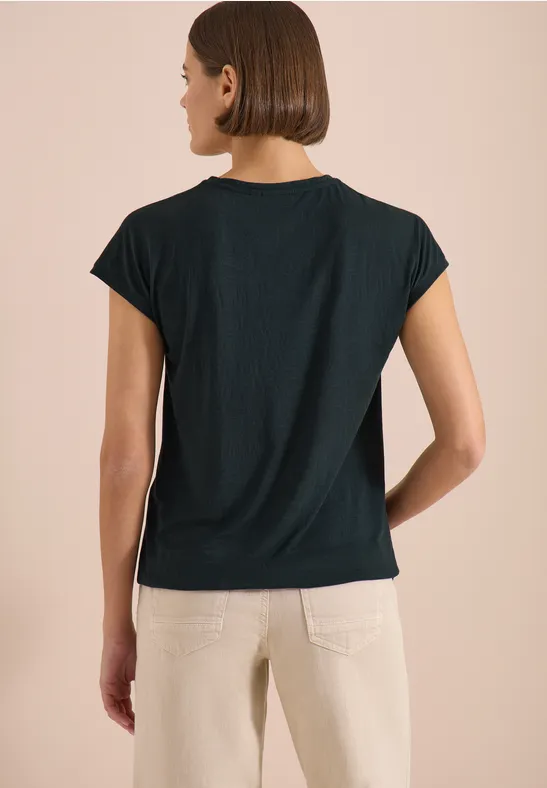 Frau steht Rücken zur Kamera, trägt schwarzes T-Shirt und helle Hose vor beigem Hintergrund.