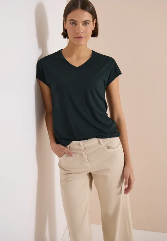 Frau lehnt entspannt an heller Wand, trägt schwarzes T-Shirt und beige Hose.