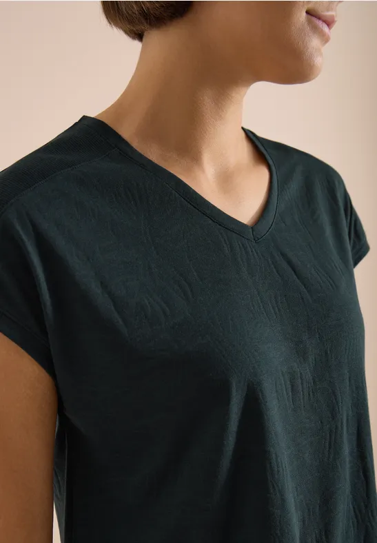 Frau trägt dunkelgrünes V-Ausschnitt-T-Shirt, Oberkörper seitlich zu sehen.
