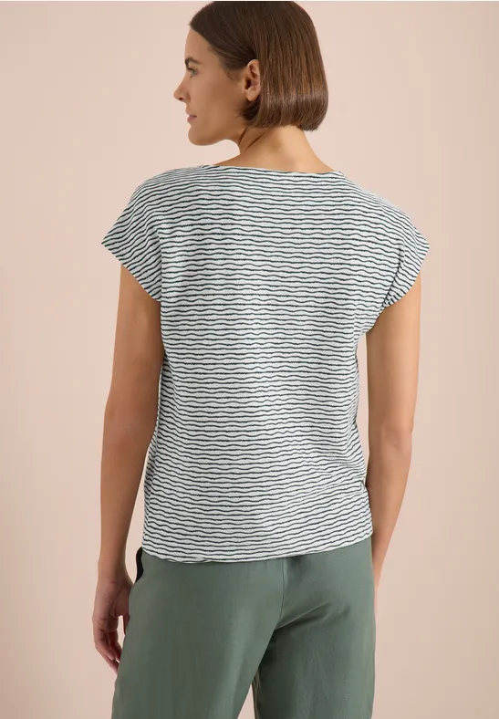 Femme de dos en t-shirt à rayures et pantalon vert devant un fond clair.