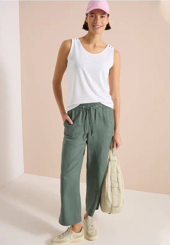 Frau trägt weißes Tanktop, locker sitzende olivgrüne Hose, weiße Sneaker und hält eine helle Stofftasche.