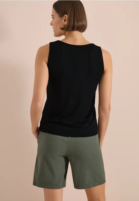 Vue de dos d’une personne portant un débardeur noir et un short vert olive sur fond beige.