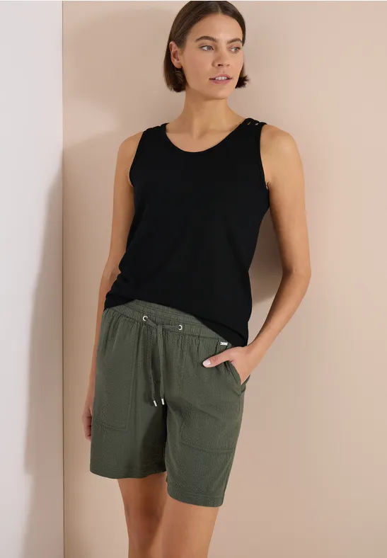 Une personne porte un débardeur noir et un short vert kaki devant un fond clair.