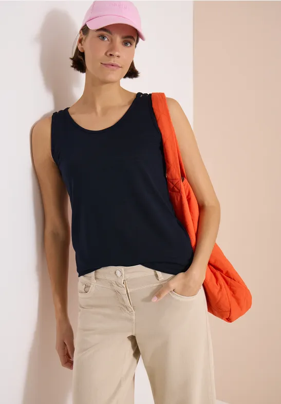 Femme appuyée contre un mur clair, porte un débardeur noir, pantalon beige, casquette rose et sac orange.