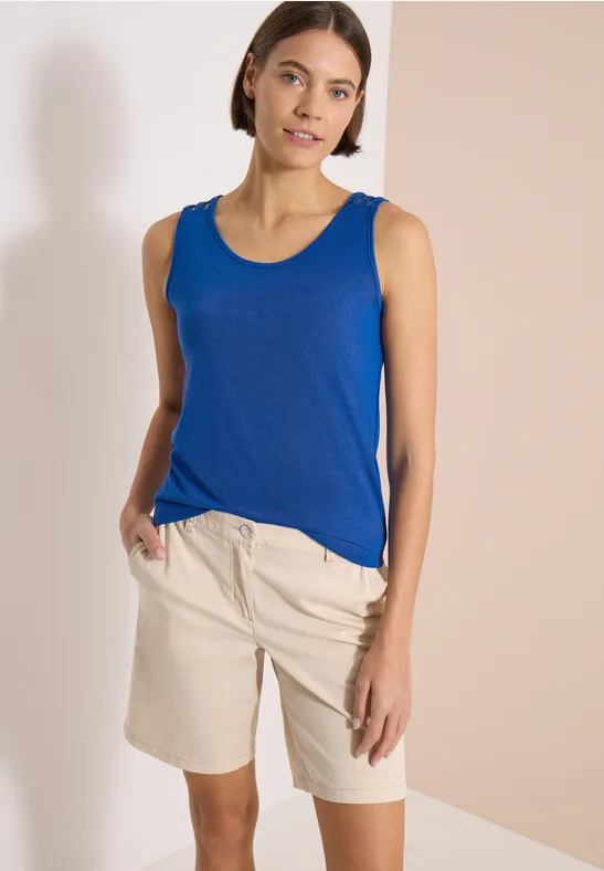 Frau trägt blaues Tanktop und beige Shorts vor cremefarbener Wand.