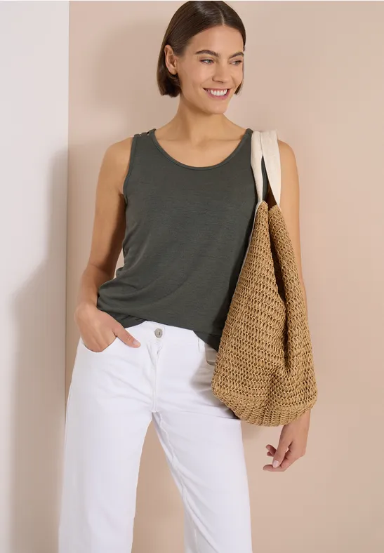 Femme en débardeur gris foncé, jean blanc et grand sac en paille, sur fond beige.