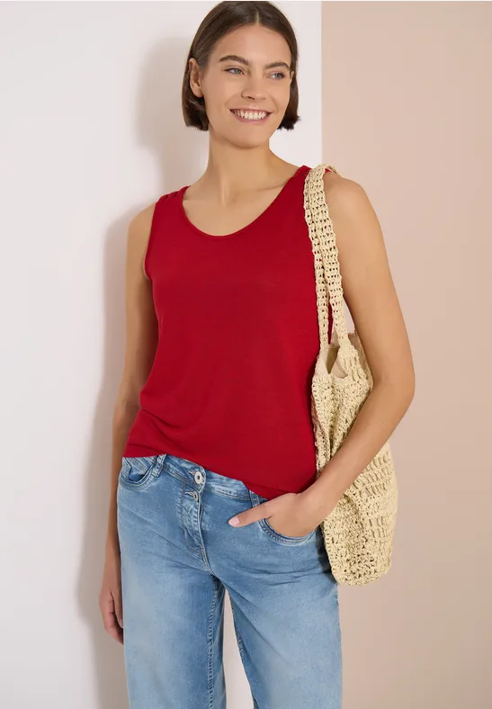 Femme en débardeur rouge, jean bleu et sac en paille devant un fond neutre.
