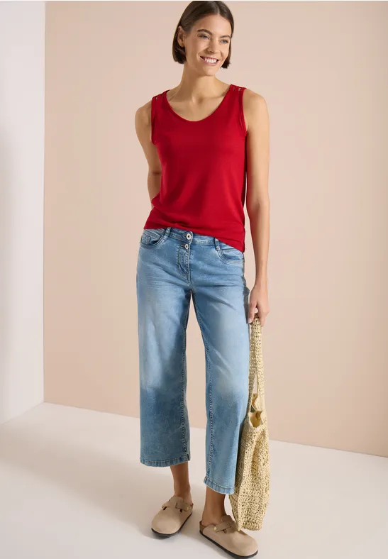 Femme portant un débardeur rouge, jean large court bleu, sac en paille et mocassins beiges devant un fond neutre.